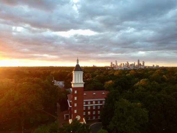 A red-brick building with a white steeple stands among green trees at sunset, with a city skyline visible in the distance under a dramatic cloudy sky.