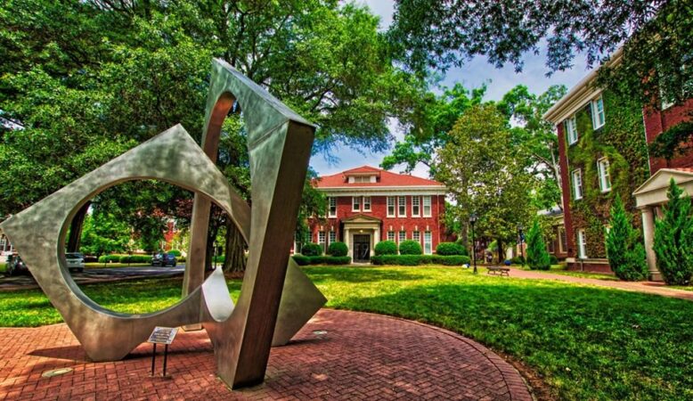 A large, abstract silver sculpture stands on a brick walkway in a green, tree-filled campus courtyard, surrounded by historic red-brick academic buildings under a partly cloudy sky.