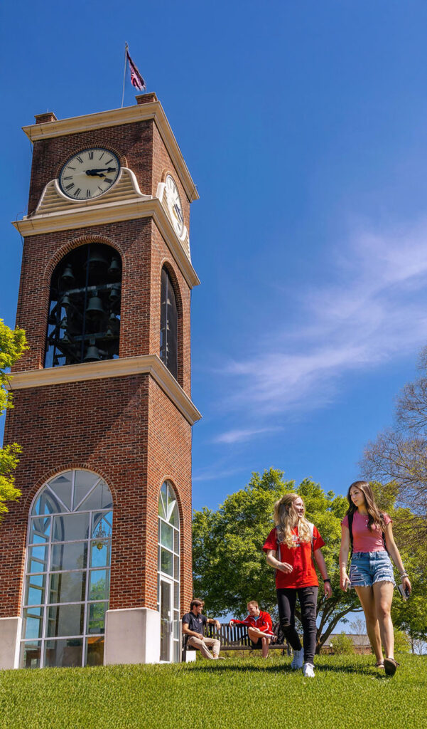 Two young women walk on grass near a tall brick clock tower under a bright blue sky. Several people sit on a bench in the background surrounded by green trees.