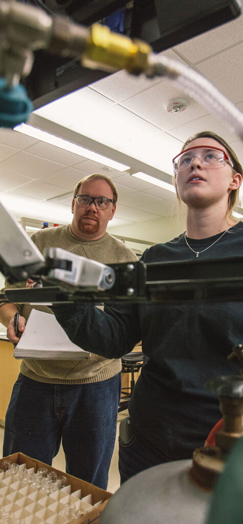 A young woman wearing safety glasses works with laboratory equipment while an older man stands nearby, observing and taking notes in a classroom or lab setting.