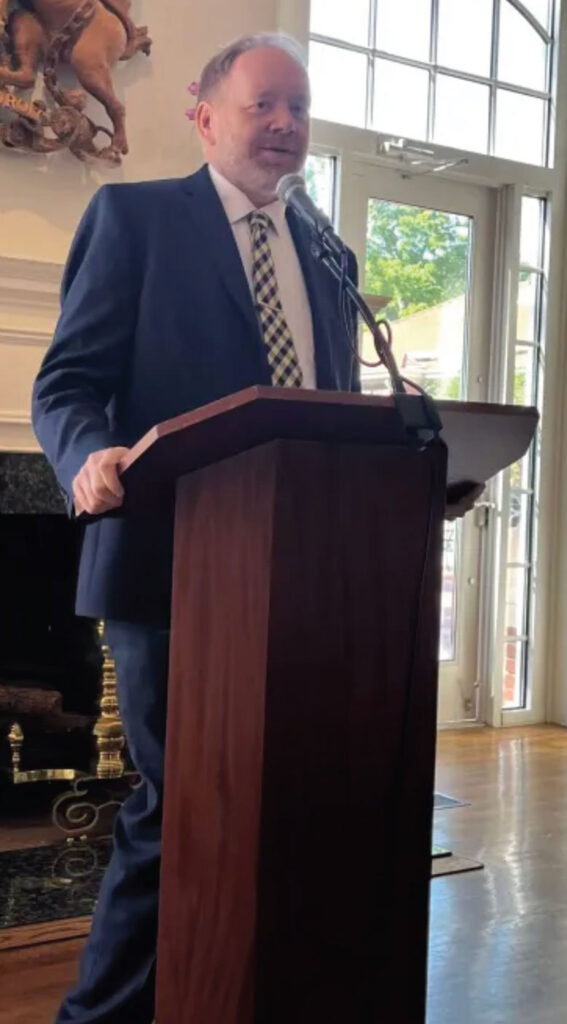 A man in a suit and checkered tie speaks at a podium with a microphone in a well-lit room with large windows and a fireplace in the background.