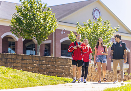 Four students walk and talk together on a sunny day outside a campus building with trees and green grass in the background.