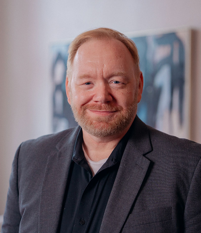 A middle-aged man with short light hair and a beard, wearing a dark blazer over a black shirt, smiles at the camera. The background is softly blurred with abstract wall art visible.