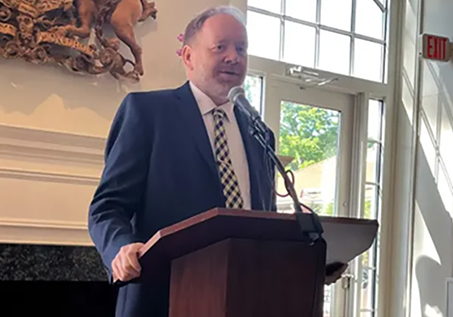 A man in a suit and checkered tie speaks at a podium in a bright room with large windows and decorative wall art behind him.