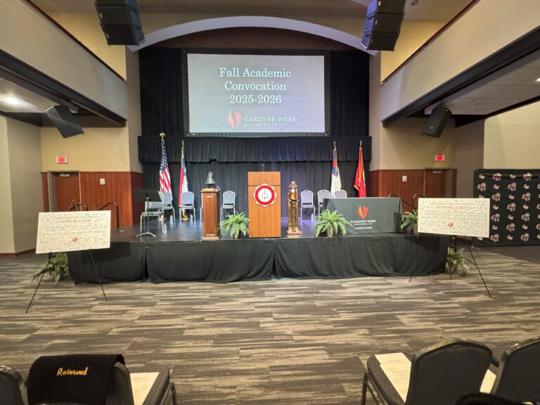 A stage set for the Fall Academic Convocation 2025-2026 at Gardner-Webb University, with podiums, flags, and two large boards with text on easels, viewed from the audience seating area.