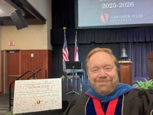 A man in academic regalia smiles for a selfie on stage. Behind him are U.S. and North Carolina flags, a large Gardner-Webb University sign, and a poster board with the university’s honor code and signatures.