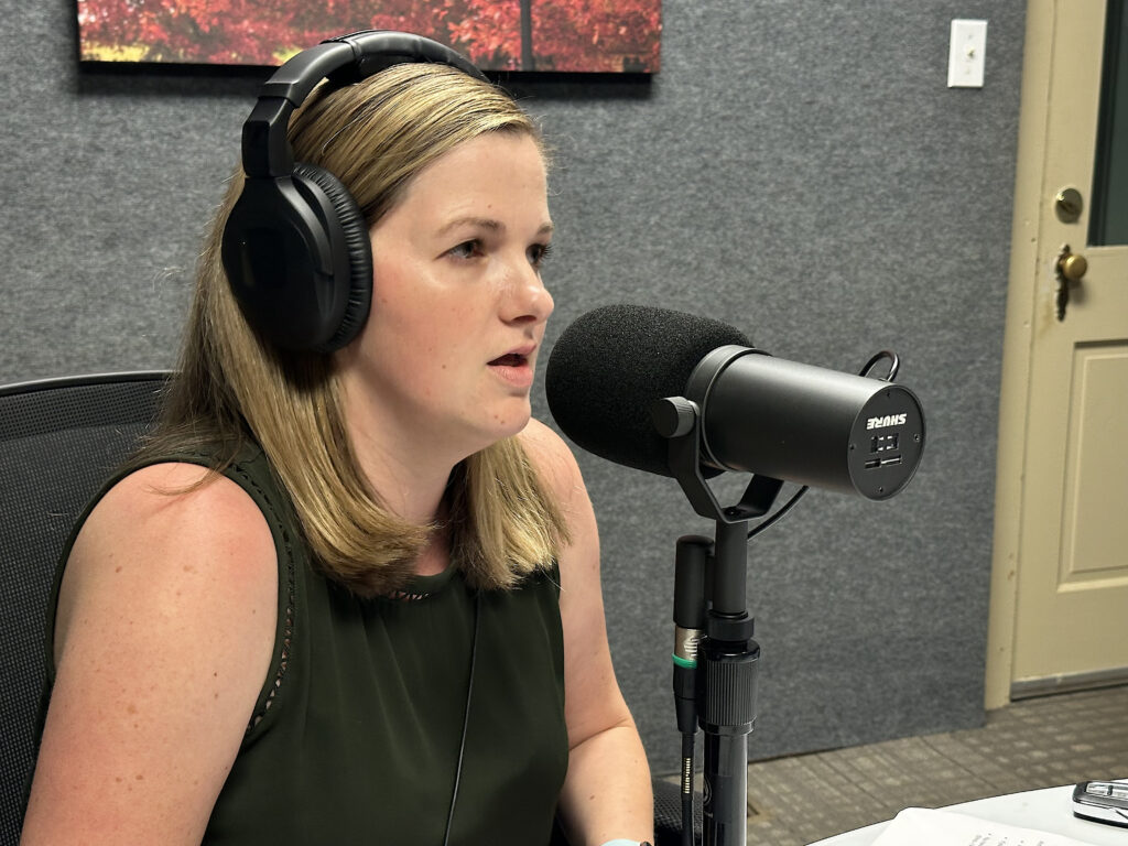 A woman with long blonde hair wearing headphones speaks into a large microphone in a recording studio. She is seated at a desk with papers in front of her and appears to be recording or broadcasting.