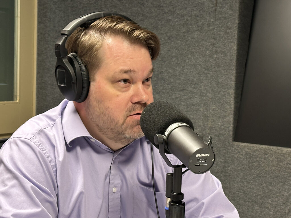 A man wearing headphones and a light purple shirt speaks into a large microphone in a recording studio with gray acoustic walls.