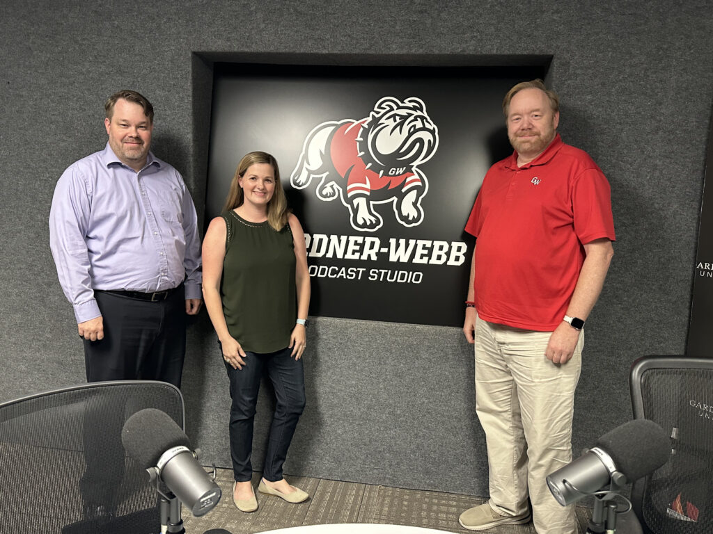 Three people stand and smile in front of a black wall sign that reads “Gardner-Webb Podcast Studio” with a bulldog mascot logo. Two men are on either side of a woman. Microphones are visible in the foreground.