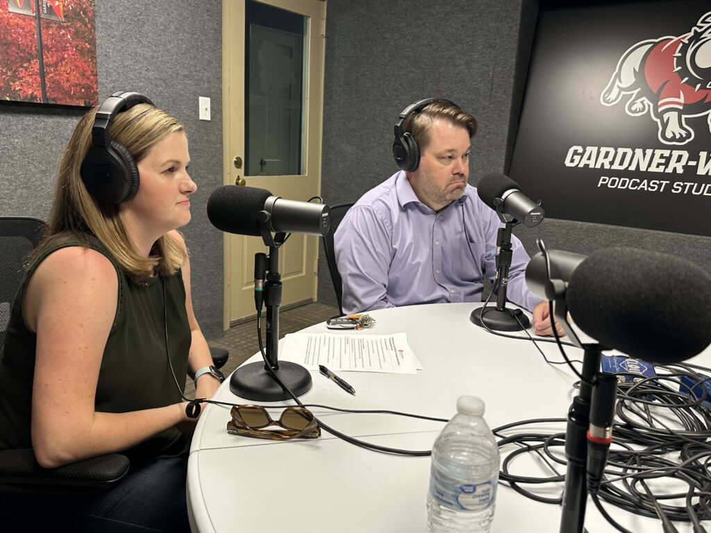 Two people wearing headphones sit at a table in a podcast studio with microphones. Papers, sunglasses, and a water bottle are on the table. The wall behind them displays the Gardner-Webb Podcast Studio logo.