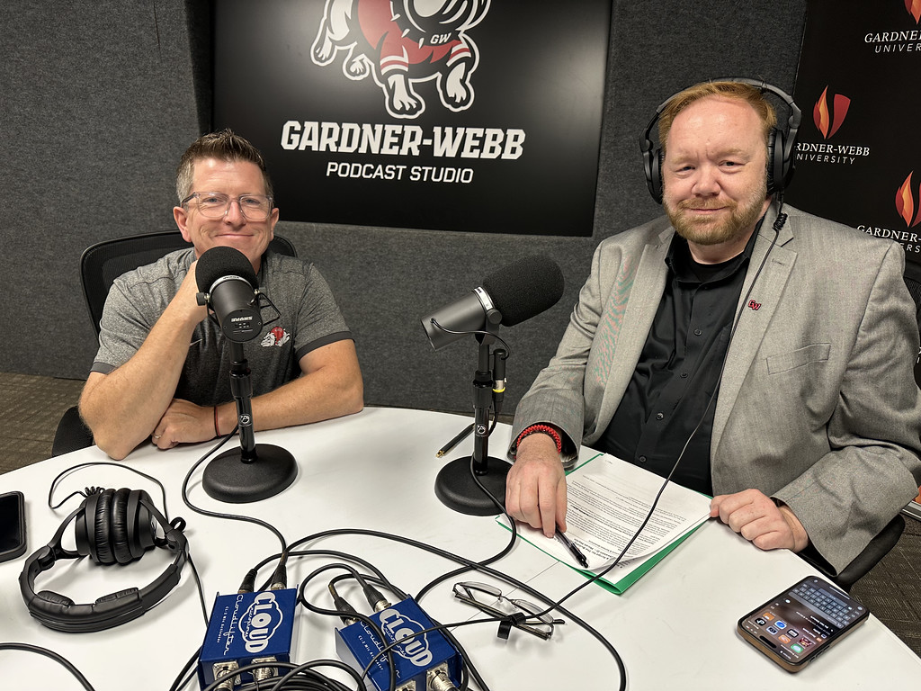 Two men sit at a podcast studio table with microphones, headphones, and equipment. A Gardner-Webb Podcast Studio sign is on the wall behind them, and both are smiling at the camera.