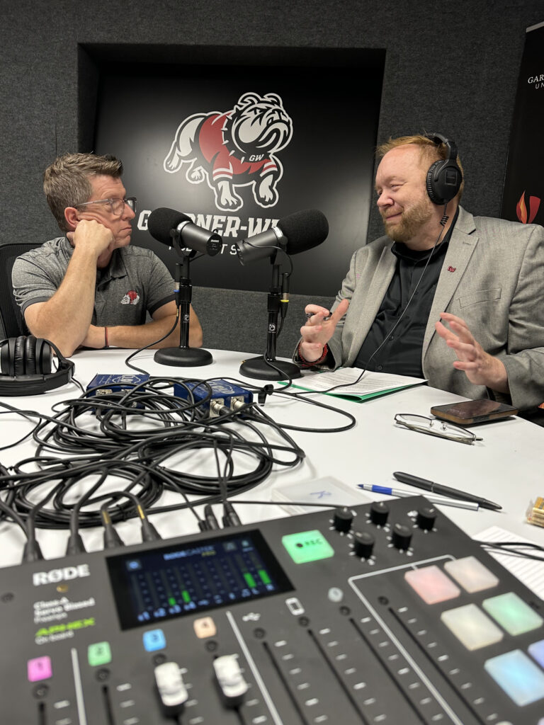 Two men sit across from each other in a recording studio, wearing headphones and speaking into microphones. Audio equipment, cables, and a notepad are visible on the table. A bulldog logo is on the wall behind them.