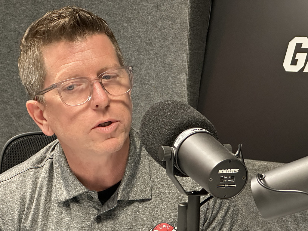 A man with short hair and glasses speaks into a black Shure microphone in a recording studio, wearing a gray polo shirt.