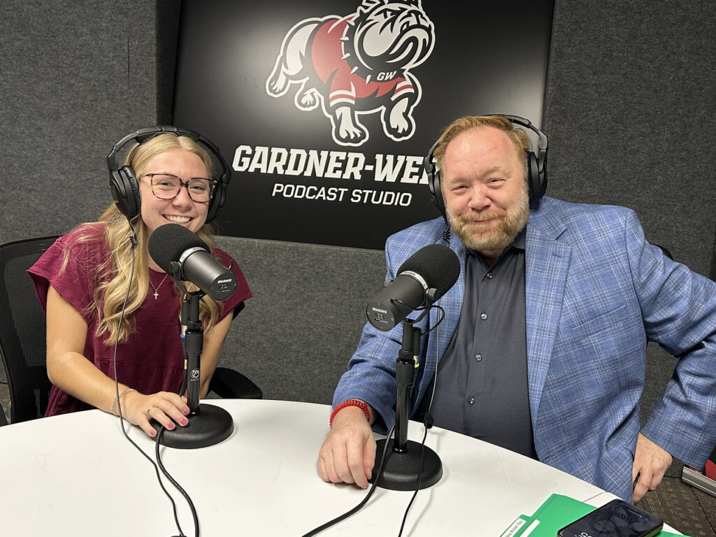 Two people wearing headphones sit at a table with microphones in a podcast studio. A Gardner-Webb University bulldog logo is visible on the wall behind them. The woman is smiling, and the man wears a blue blazer and smiles slightly.