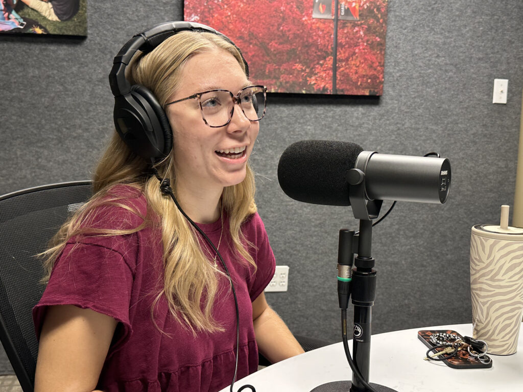 A young woman with long blonde hair and glasses, wearing a maroon shirt and large headphones, speaks into a microphone in a recording studio. A tumbler and keys sit on the table in front of her.