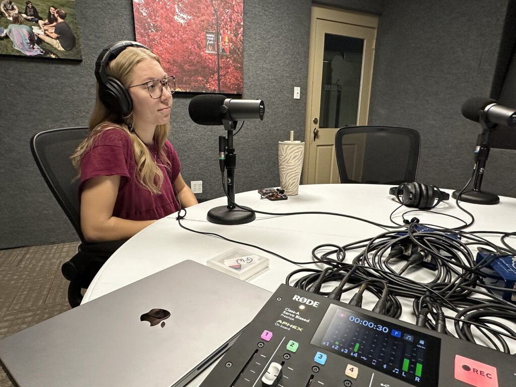 A woman wearing headphones speaks into a microphone in a podcast recording studio. A mixer, headphones, laptop, and drink cup are on the round table. Framed photos hang on the gray walls in the background.
