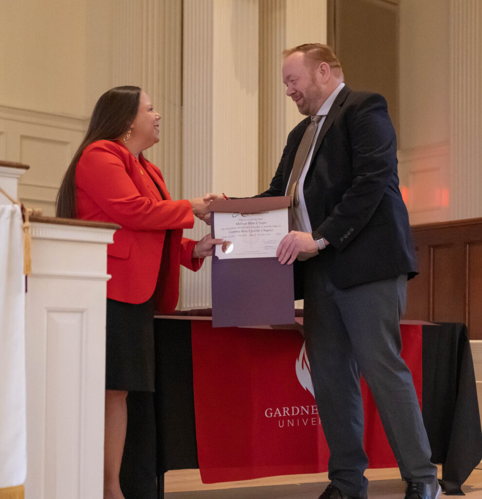 A woman in a red blazer hands a certificate to a smiling man in a suit during a ceremony. They stand in front of a red tablecloth with a Gardner-Webb University logo in a formal room with columns.