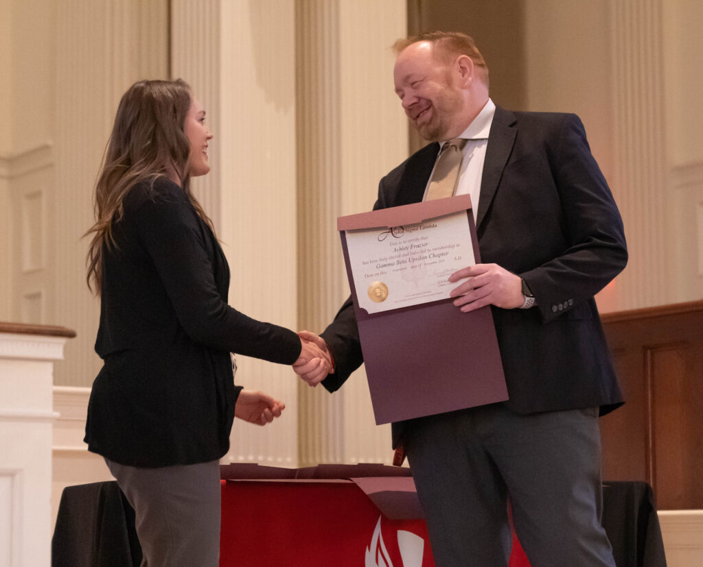 A man in a suit hands a woman an award certificate while they shake hands and smile, standing in a formal indoor setting with columns and a table in the background.