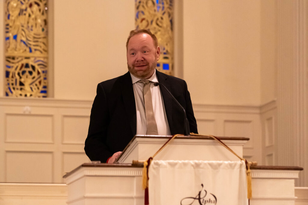 A man in a suit and tie stands at a podium, speaking in what appears to be a church or formal setting, with ornate gold decorations and stained-glass windows in the background.