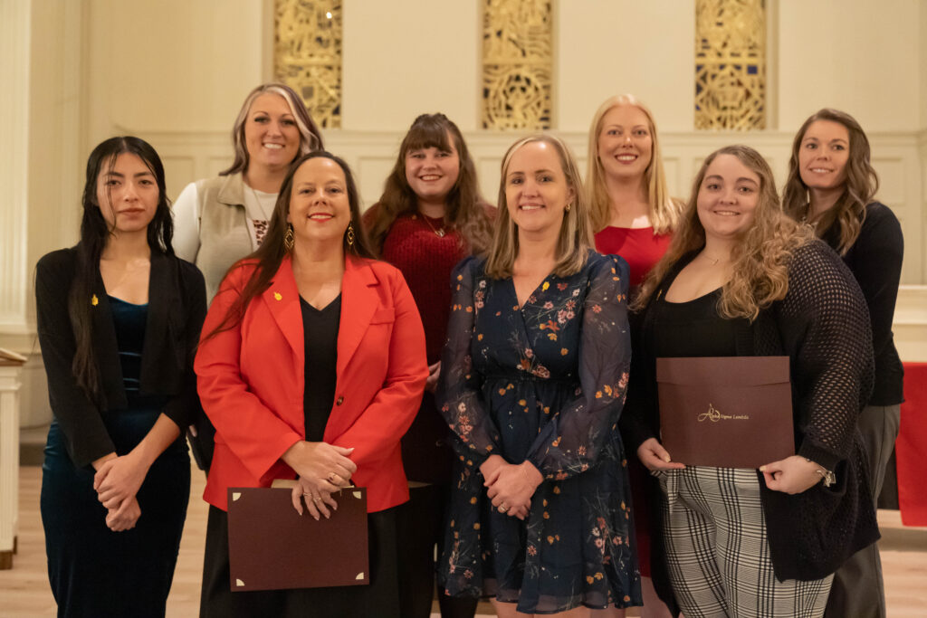 Eight women stand together indoors, some holding certificates. They are dressed in business or semi-formal attire and smiling at the camera. The background features ornate panels and cream-colored walls, suggesting a formal event or ceremony.