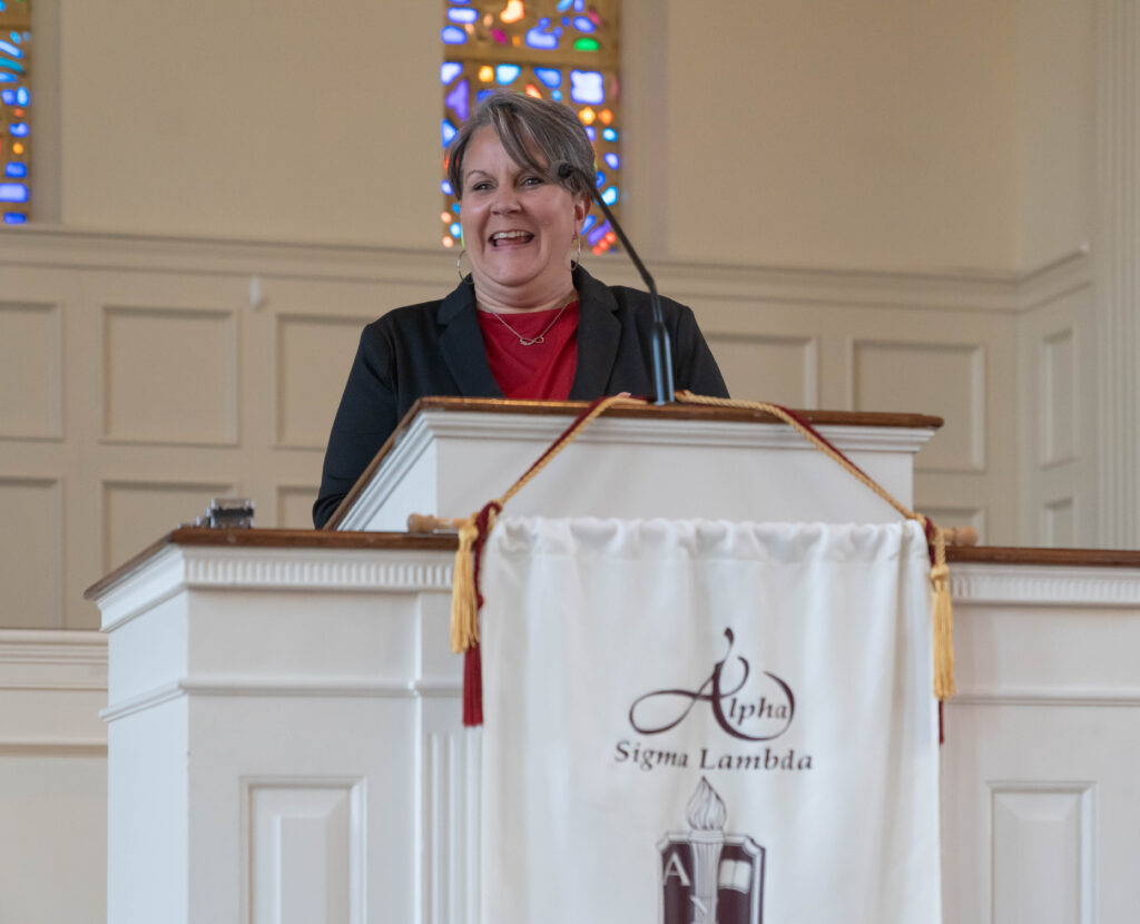 A woman in a black jacket stands at a white podium with an Alpha Sigma Lambda banner, speaking in a church-like setting with stained glass windows in the background.