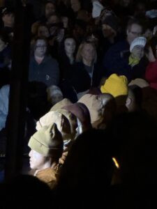 A group of people wearing tan and brown hats stand in front of a crowd at night, with some faces illuminated by light while others remain in shadow. The audience watches attentively in the background.
