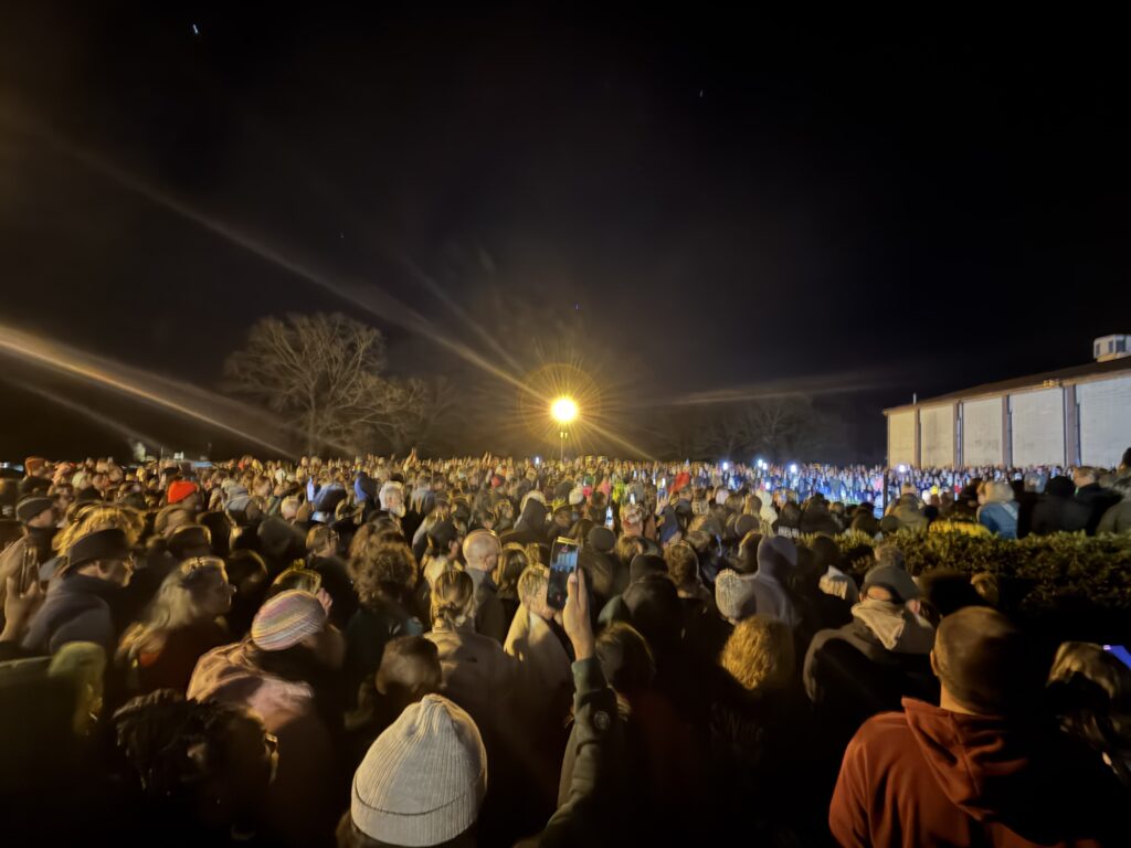 A large crowd gathered outdoors at night, illuminated by a bright light in the background. Many people are wearing winter clothing, and some are holding up phones to capture the scene.