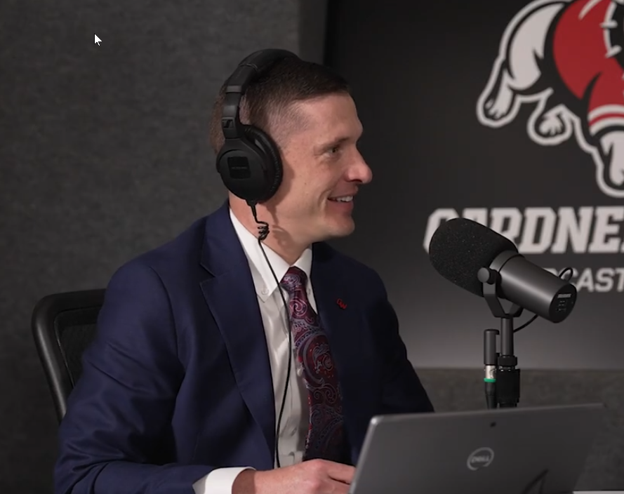 A man in a navy suit, patterned tie, and headphones speaks into a microphone in a podcast studio with a laptop in front of him. The background features part of a bulldog logo and the word CARDINALS.