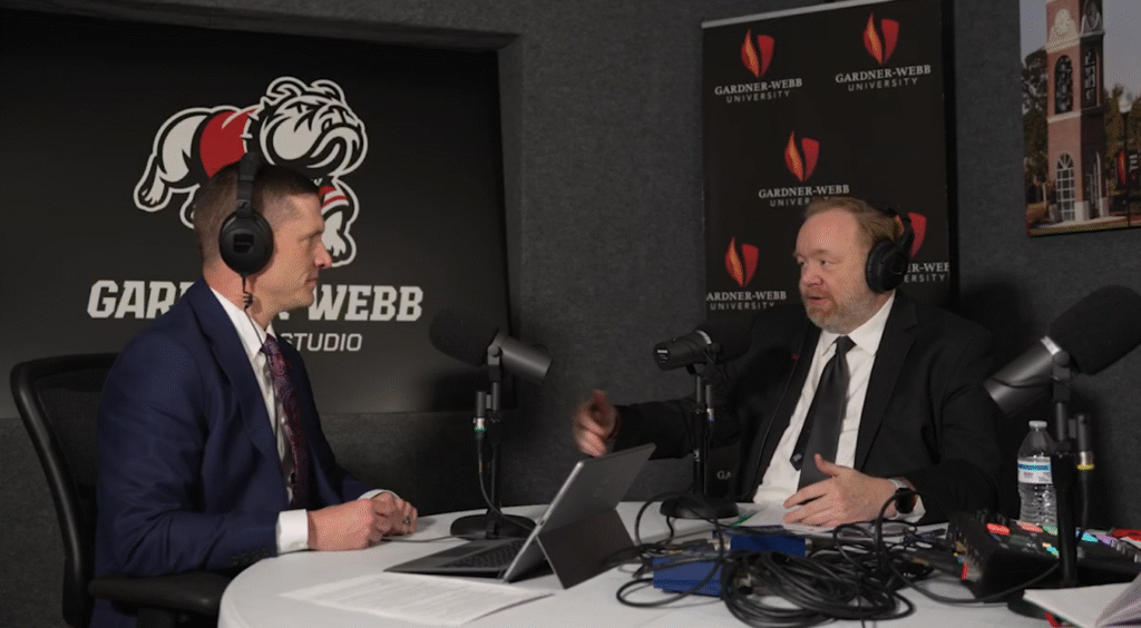 Two men in suits sit at a round table with microphones and laptops, recording a podcast in a studio featuring Gardner-Webb University logos and a large bulldog mascot graphic on the wall.