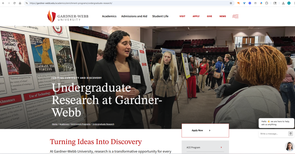 A woman presents her research poster to attendees at an academic event in a large hall. Other students and posters are visible in the background. The banner reads, “Undergraduate Research at Gardner-Webb.”.