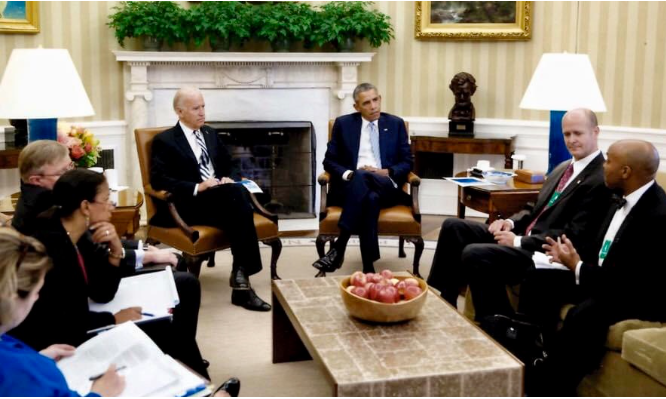 A group of people sit in a formal meeting in the Oval Office, with two men in suits seated near the fireplace, and others around them taking notes. A coffee table with a bowl of apples is in the center.
