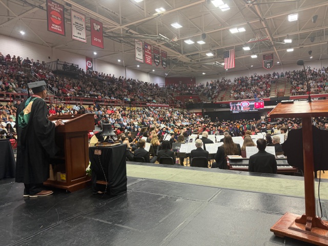 A large graduation ceremony in a gymnasium, with a graduate speaking at a podium, an audience filling the bleachers, and a band or orchestra seated onstage. Banners and an American flag hang above.