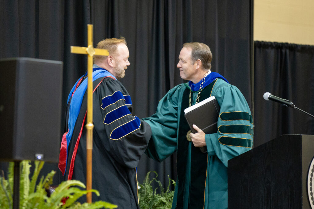 Two men in academic regalia shake hands at a graduation ceremony, standing next to a podium and a gold cross, with black curtains and greenery in the background.