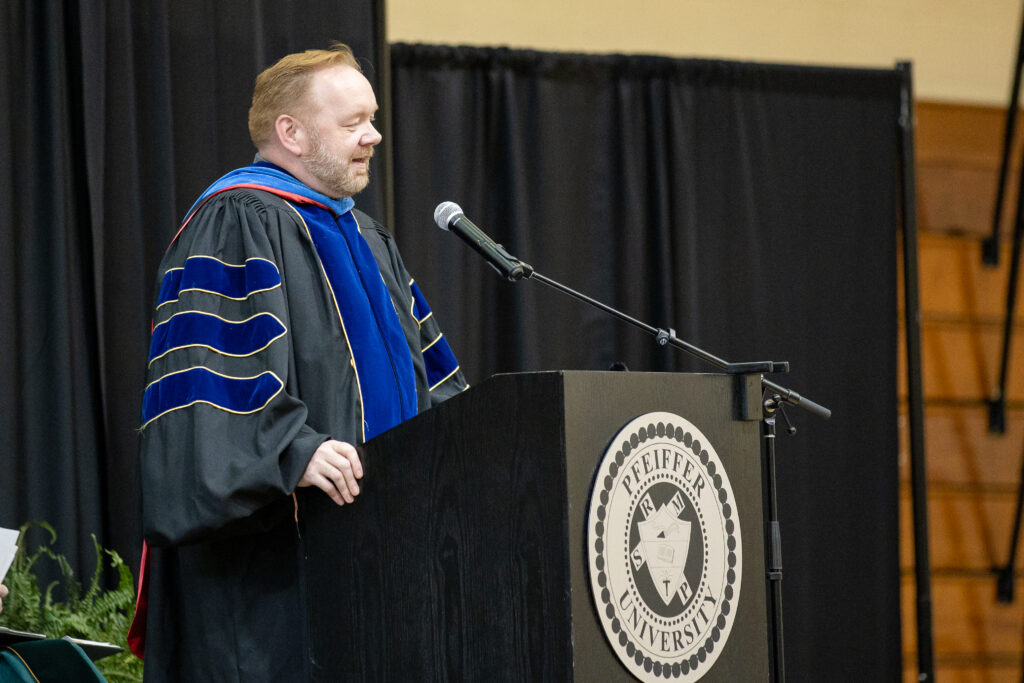 A person in academic regalia speaks at a podium with a Pfeiffer University seal, during an indoor event.